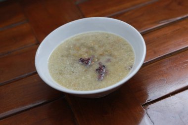 a close up of a bowl of green bean porridge and black sticky rice doused in coconut milk, served warm and ready to eat on a wooden background with a natural pattern. Indonesian food concept.