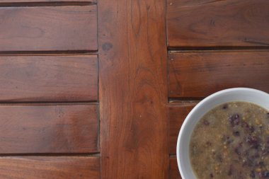 a close up of a bowl of green bean porridge and black sticky rice doused in coconut milk, served warm and ready to eat on a wooden background with a natural pattern. Indonesian food concept.