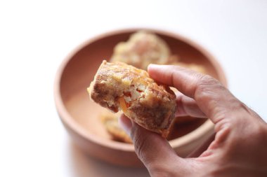 a close up of fried stuffed tofu or Gorengan Tahu Isi holding in hand isolated on white background and bowl of stuffed fried tofu blurry. Indonesian food concept.