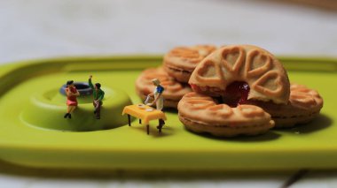 Miniature figures of a couple talking on a green dining table near a chef cooking on a background of a cake filled with strawberry jam. discussion concept.