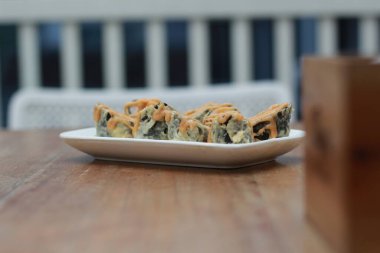 Japanese food called Volcano Salmon Roll Sushi. Served warm on a white rectangular plate on the table with a slanted cut.