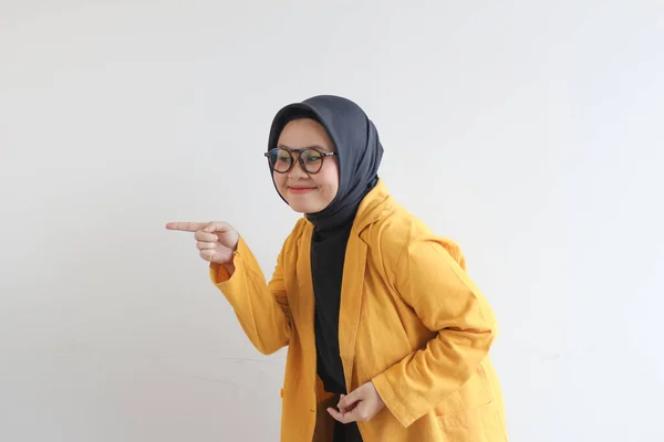年轻美丽的亚洲穆斯林女子 戴着眼镜 头戴头巾 身穿黄色夹克 满面春风 两只手都带着自信而快乐的微笑 两只手指向被白色背景隔离的侧面 广告理念 — 图库照片
