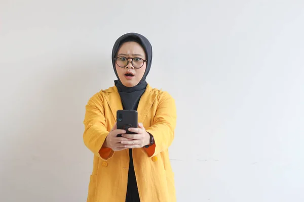 眼鏡ヒジャーブ州の美しい若いアジアの女性と黄色のブレザーを身に着けている彼女の携帯電話を見てショックを受けた顔白い背景に隔離された — ストック写真