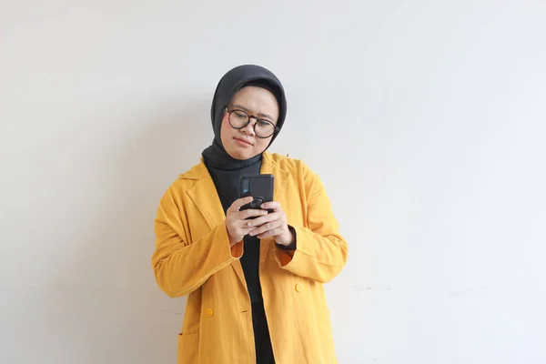 年轻美丽的亚洲女人 戴着眼镜 头戴头巾 头戴黄色的夹克 看着她的手机 她的脸直挺挺的 背景是白色的 — 图库照片