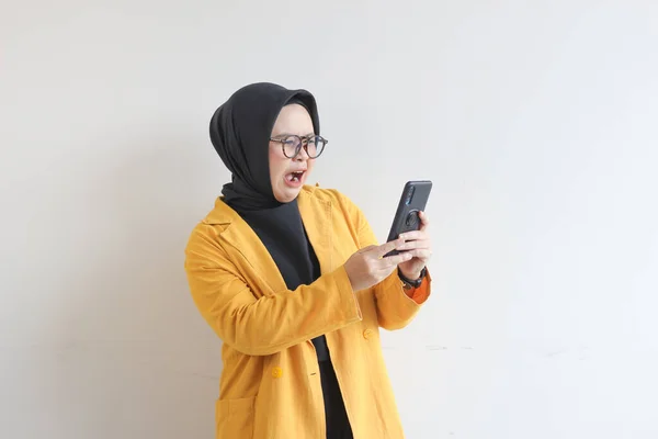 年轻美丽的亚洲女人 戴着眼镜 头戴头巾 身穿黄色夹克 看着她的手机 脸上带着震惊的表情 背景是白色的 — 图库照片