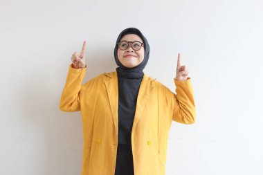 Gözlüklü, tesettürlü, sarı ceketli, kendinden emin ve mutlu bir şekilde gülümseyen, her iki eli de beyaz arka planda izole edilmiş, reklamcı bir kadın.