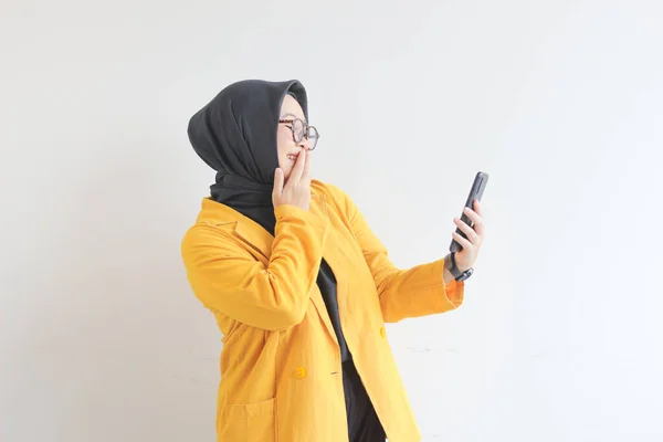 眼鏡ヒジャーブ州の美しい若いアジアの女性と黄色のブレザーを身に着けている彼女の携帯電話を見て幸せな顔白い背景に隔離された — ストック写真