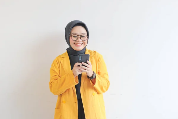 眼鏡ヒジャーブ州の美しい若いアジアの女性と白い背景に隔離された幸せな顔で彼女の携帯電話を見て黄色のブレザーを身に着けて — ストック写真