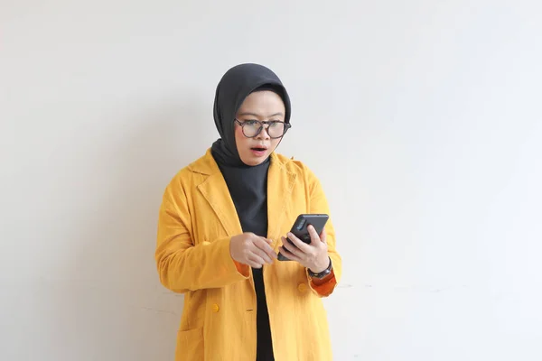 眼鏡ヒジャーブ州の美しい若いアジアの女性と黄色のブレザーを身に着けている彼女の携帯電話を見てショックを受けた顔白い背景に隔離された — ストック写真