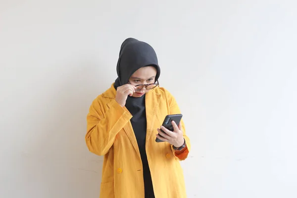 年轻美丽的亚洲女人 戴着眼镜 头戴头巾 身穿黄色夹克 看着她的手机 脸上带着震惊的表情 背景是白色的 — 图库照片