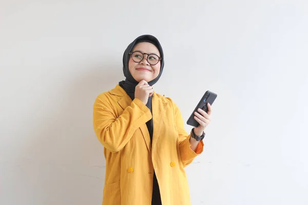 Beautiful young Asian Muslim woman, wearing glasses and yellow blazer looking sideways while smiling and holding mobile phone isolated on white background.