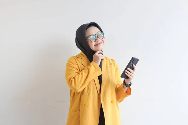 美しい若いアジアのイスラム教徒の女性 笑顔と白い背景に隔離された携帯電話を保持しながら横に見える眼鏡と黄色のブレザーを身に着けています — ストック写真