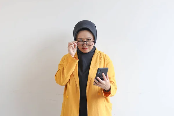 年轻美丽的亚洲女人 戴着眼镜 头戴头巾 身穿黄色夹克 面带震惊地看着相机 手持白色背景的手机 — 图库照片