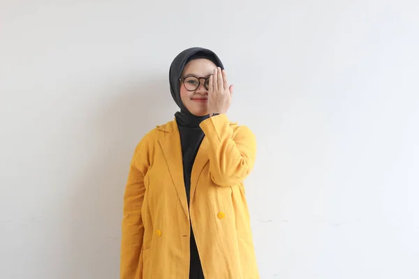 年轻美丽的亚洲穆斯林妇女 戴着眼镜 头戴黄色的夹克 手捂住脸的一侧 面带白色背景 面带微笑 — 图库照片