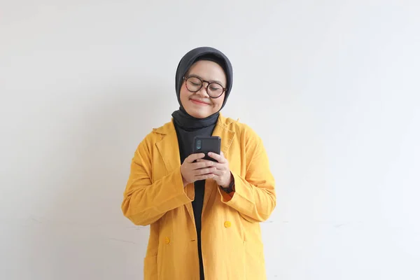 年轻美丽的亚洲女人 戴着眼镜 头戴头巾 头戴黄色的夹克 看着她的手机 脸上挂着幸福的脸 背景是白色的 — 图库照片