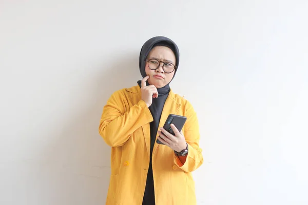 年轻美丽的亚洲穆斯林妇女 戴着眼镜 身穿黄色夹克 一边侧看一边拿着白色背景的手机 — 图库照片