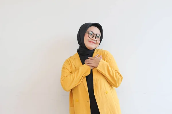 年轻美丽的亚洲穆斯林妇女 戴着眼镜 身穿黄色夹克 微笑着 双手放在胸前 露出深情的姿态 与白色背景格格不入 — 图库照片