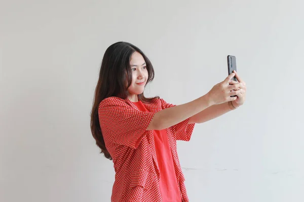 照片上美丽的亚洲女人穿着红色衣服 看着手机 脸上挂着快乐的表情 背景是白色的 — 图库照片