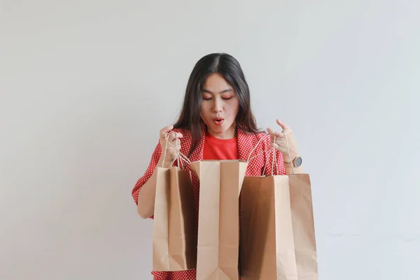 ショッピングバッグのコンテンツを見ながら驚いたジェスチャーで赤い服を着た美しいアジアの女性の肖像画 隔離された白い背景 — ストック写真