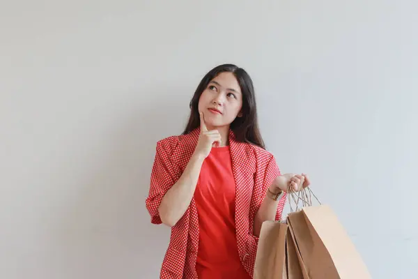 白い背景に隔離された思考表現で多くのショッピングバッグを運ぶ赤い服を身に着けている美しいアジアの女性の肖像画 — ストック写真