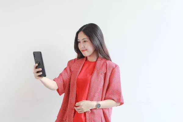 照片上美丽的亚洲女人穿着红色衣服 看着手机 脸上挂着快乐的表情 背景是白色的 — 图库照片