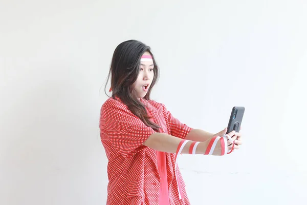 在印尼独立日这天 身穿红色服装的美丽的亚洲女人的肖像 看着手机 脸上带着惊讶的表情 与白色背景隔离 — 图库照片