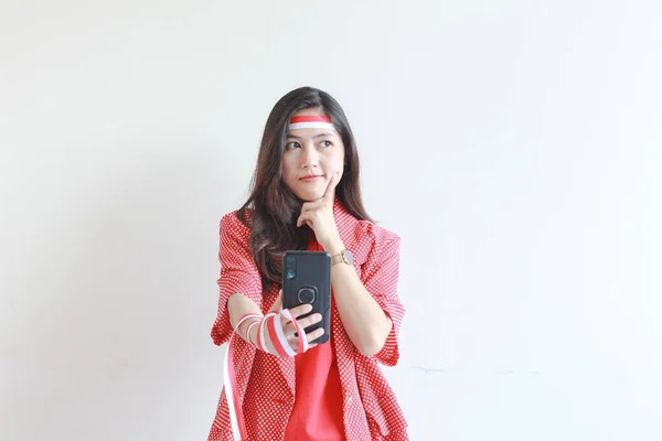 インドネシア独立記念日を祝う赤い衣装を着た美しいアジアの女性の肖像画は 白い背景に孤立した思考表現で携帯電話を保持しています — ストック写真