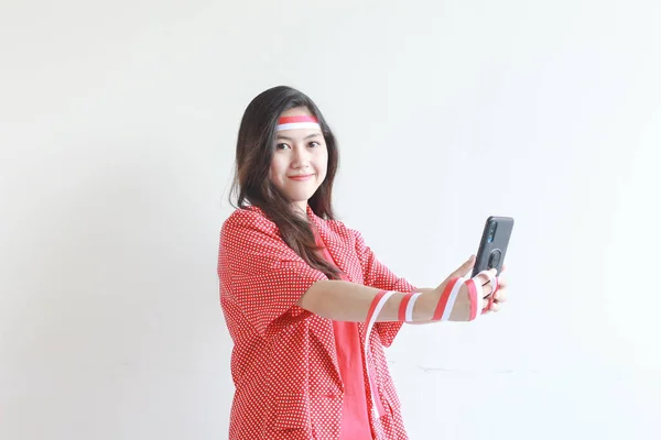 インドネシアの独立記念日を祝う赤い衣装を着た美しいアジアの女性の肖像画 携帯電話を保持し 白い背景で孤立した笑顔 — ストック写真