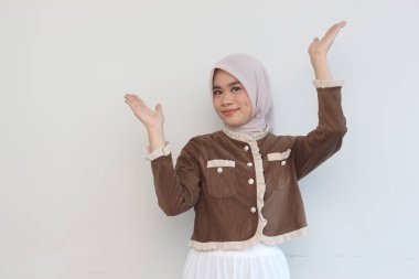 A young woman wearing a brown corduroy jacket and hijab presents with open palms against a white background. She is smiling and appears relaxed and cheerful. clipart