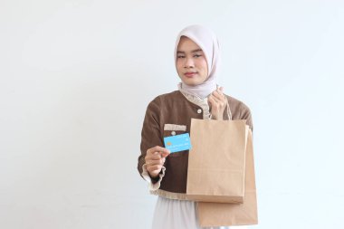 Başörtüsü takmış genç bir kadın, elinde alışveriş poşetleri ve kredi kartı ile kendinden emin bir şekilde gülümsüyor. Çevrimiçi alışveriş başarılı.