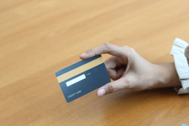 Tahta bir masada kredi kartını tutan bir elin yakın çekimi. Kart koyu mavi ve altın çizgili..