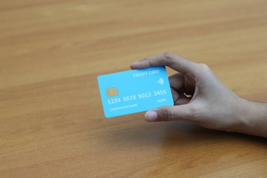 Ahşap bir masanın üzerinde açık mavi bir kredi kartı tutan bir elin yakın çekimi. Görüntü çevrimiçi alışveriş ve dijital ödemeleri sembolize ediyor.