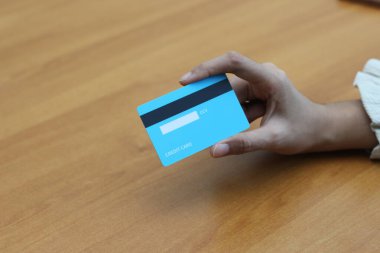 Tahta bir yüzey üzerinde açık mavi bir kredi kartı tutan bir kişi. Görüntü online alışveriş ve finansal işlemleri öneriyor.
