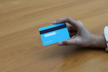 Bir el ahşap bir masada açık mavi bir kredi kartı tutuyor. Odak noktası kart ve el..