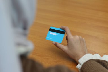 Bir kadının eli ahşap bir masada açık mavi bir kredi kartı tutuyor. Resim çevrimiçi ödeme veya aktarımı betimler.