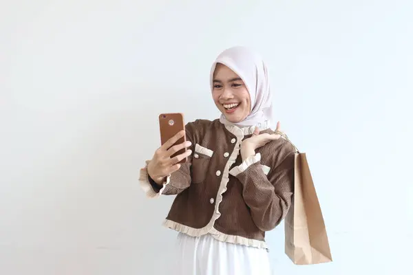 Tesettürlü neşeli bir kadın alışveriş torbası tutarken ve telefonunu kullanırken gülümsüyor. Başarılı bir online alımdan sonra mutlu görünüyor..