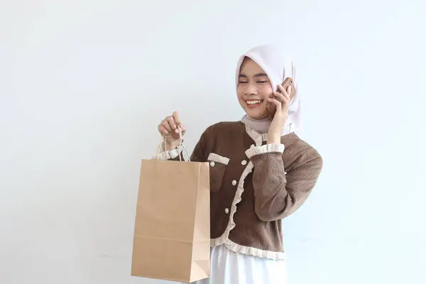 Tesettürlü neşeli bir kadın elinde alışveriş torbasıyla telefonuyla konuşuyor. Çevrimiçi alımından memnun..