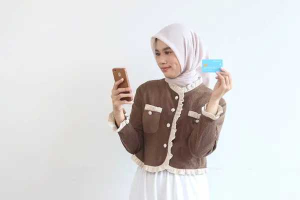 Türbanlı genç bir kadın internet üzerinden alışveriş yapmak için akıllı telefonu ve kredi kartını kullanıyor. Gülümsüyor ve mutlu görünüyor..