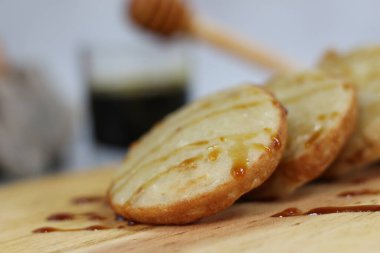 Tahtaya bal serpiştirilmiş üç mini krep yakın plan. Bulanık bir arkaplan, bal arayıcıyı ve koyu bir içeceği gösteriyor..