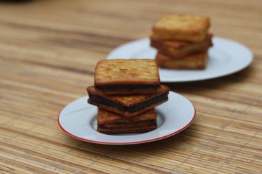 Beyaz bir tabakta bir yığın çikolatalı sandviç krakeri ahşap bir yüzeye oturur. Arka planda başka bir yığın görünür.