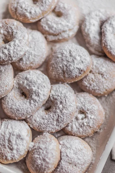 Many round donuts covered with powdered sugar on a cream enameled baking sheet.
