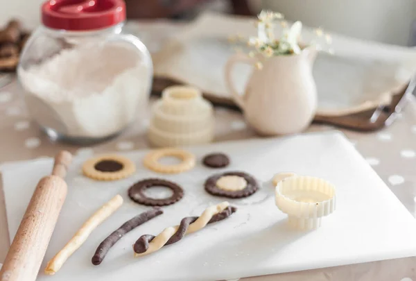 Cut from a short pastry and cooked cookies of different shapes on a cutting board, along with a can of flour, a rolling pin, a baking sheet with baking paper and a jug with a sprig of linden.