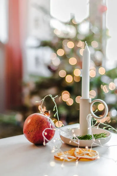 A lit candle in a ceramic candlestick on a table with pomegranate and orange slices against the background of a decorated Christmas tree.
