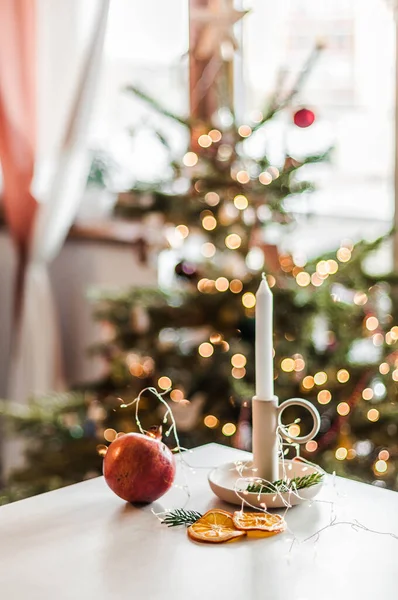A lit candle in a ceramic candlestick on a table with pomegranate and orange slices against the background of a decorated Christmas tree.