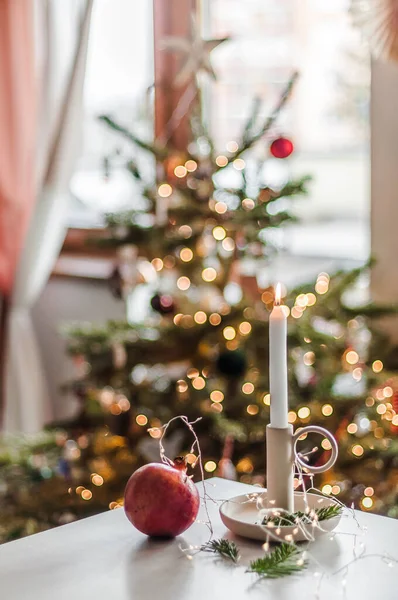 A lit candle in a ceramic candlestick on a table with pomegranate against the background of a decorated Christmas tree.