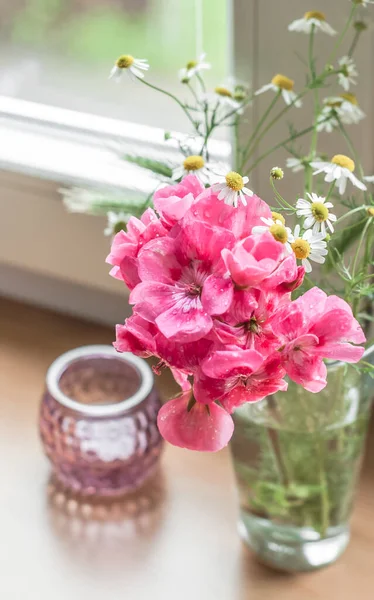 A small bouquet of poza geranium flowers, field daisies and spikelets in a glass on a windowsill against the background of green foliage outside the window and a berry-colored candlestick top view.