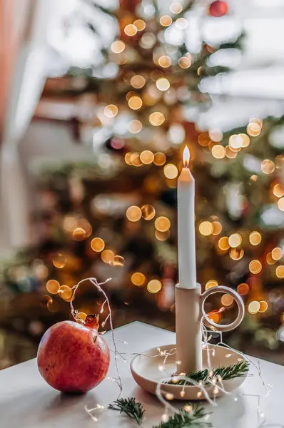 A lighted candle in a ceramic candlestick on a table with pomegranate and fir tree twigs closeup against the background of a Christmas tree.