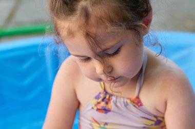 Küçük kız arka bahçedeki mavi havuzda suyla oynuyor.
