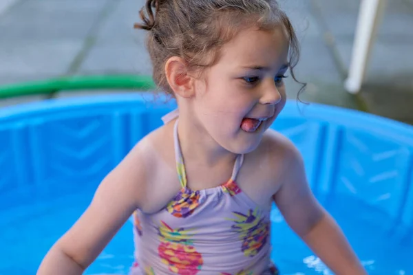 Küçük kız arka bahçedeki mavi havuzda suyla oynuyor.