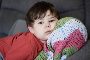 Kafası battaniyesinin içinde bir topun üzerinde kanepede dinlenen genç bir çocuğun portresi.